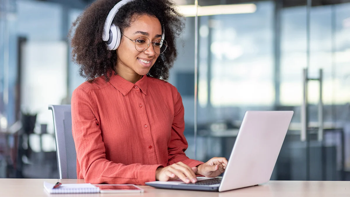 Woman looking at her laptop screen and smiling while listening to audio on her headphones.